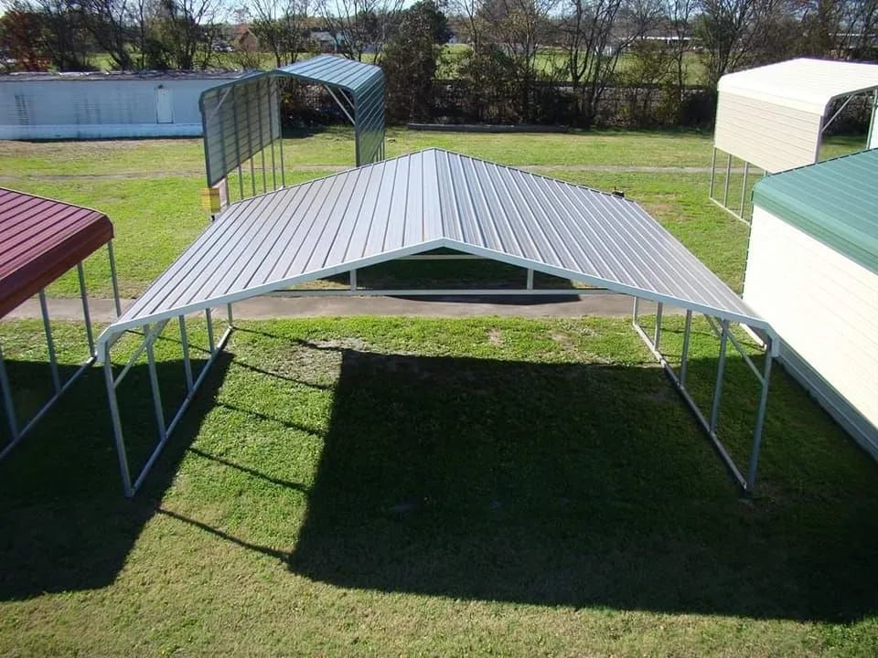 Metal carport with peaked roof on grassy lawn between several buildings