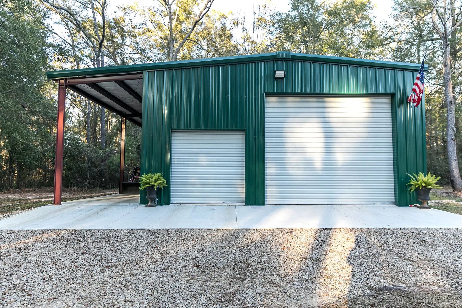 Green metal garage with two roll-up doors and American flag in wooded area